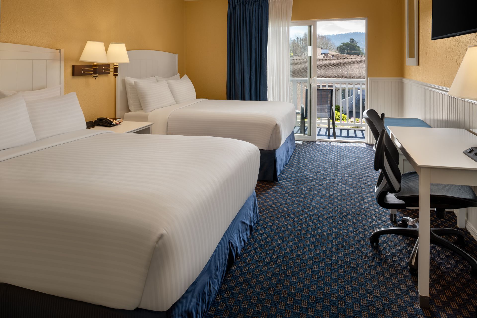 Hotel room with two beds, desk, and balcony door overlooking a mountain view