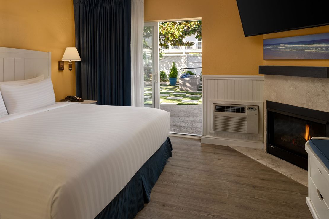 A hotel room with a white-bedded king size bed, dark curtains, a fireplace, and a glass door leading to an outdoor patio.