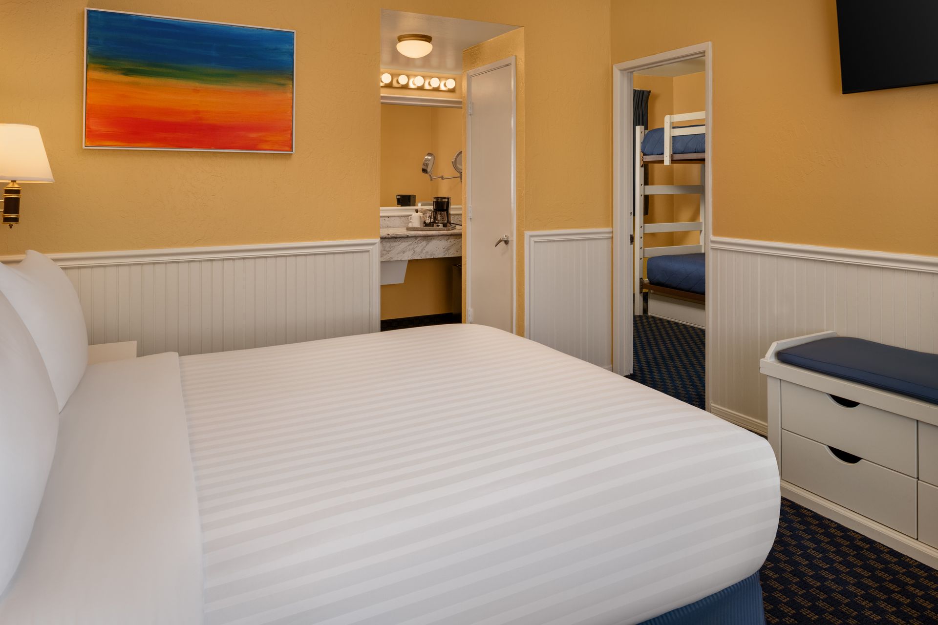 Bright bedroom with a white bed, yellow walls, colorful painting, and open bathroom and closet doors