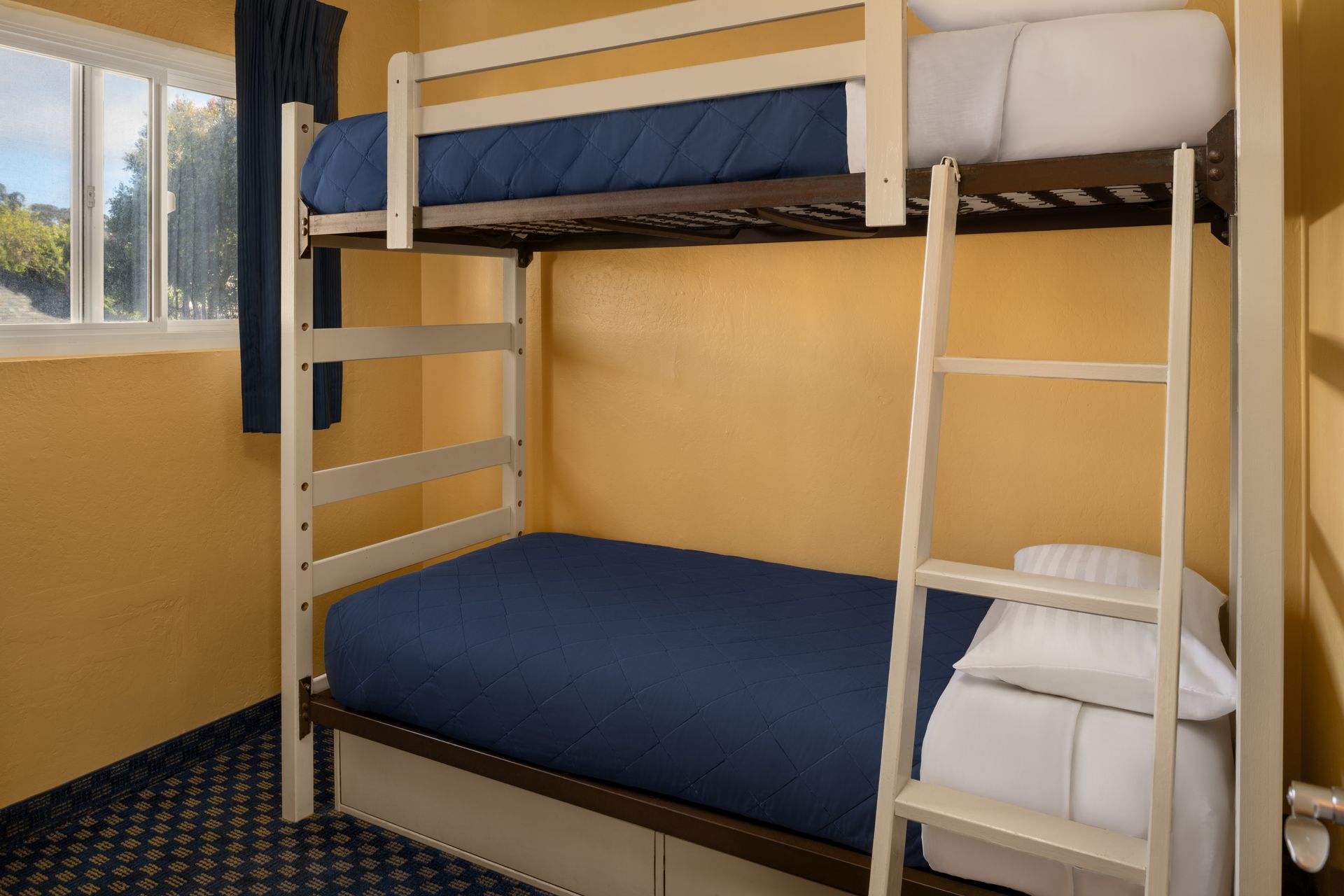 Dorm room with wooden bunk beds, blue mattresses, and a window with daylight