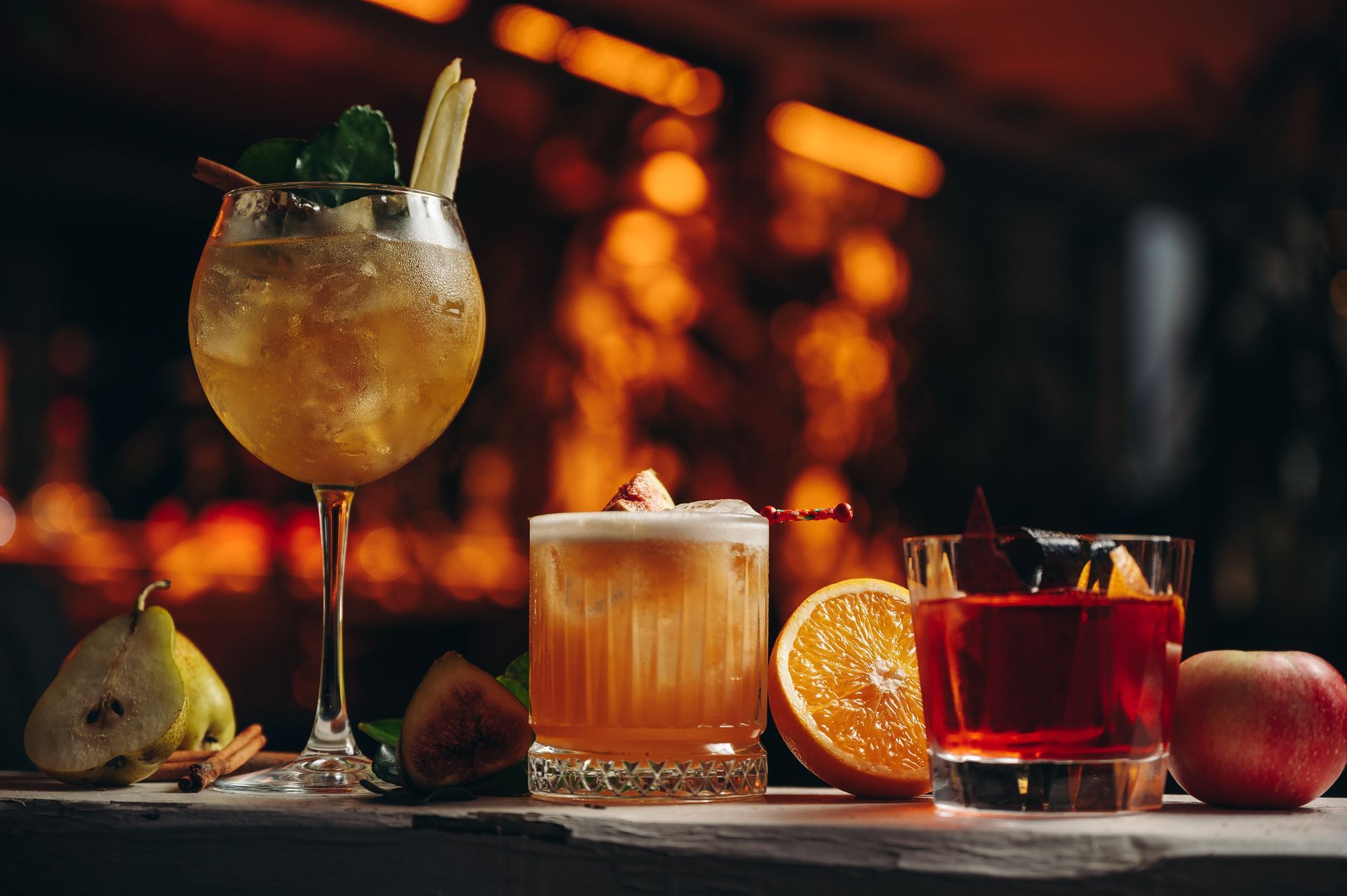 Three cocktails arranged on a wooden surface with a sliced pear, orange, and apple against a warm, blurry bar background.