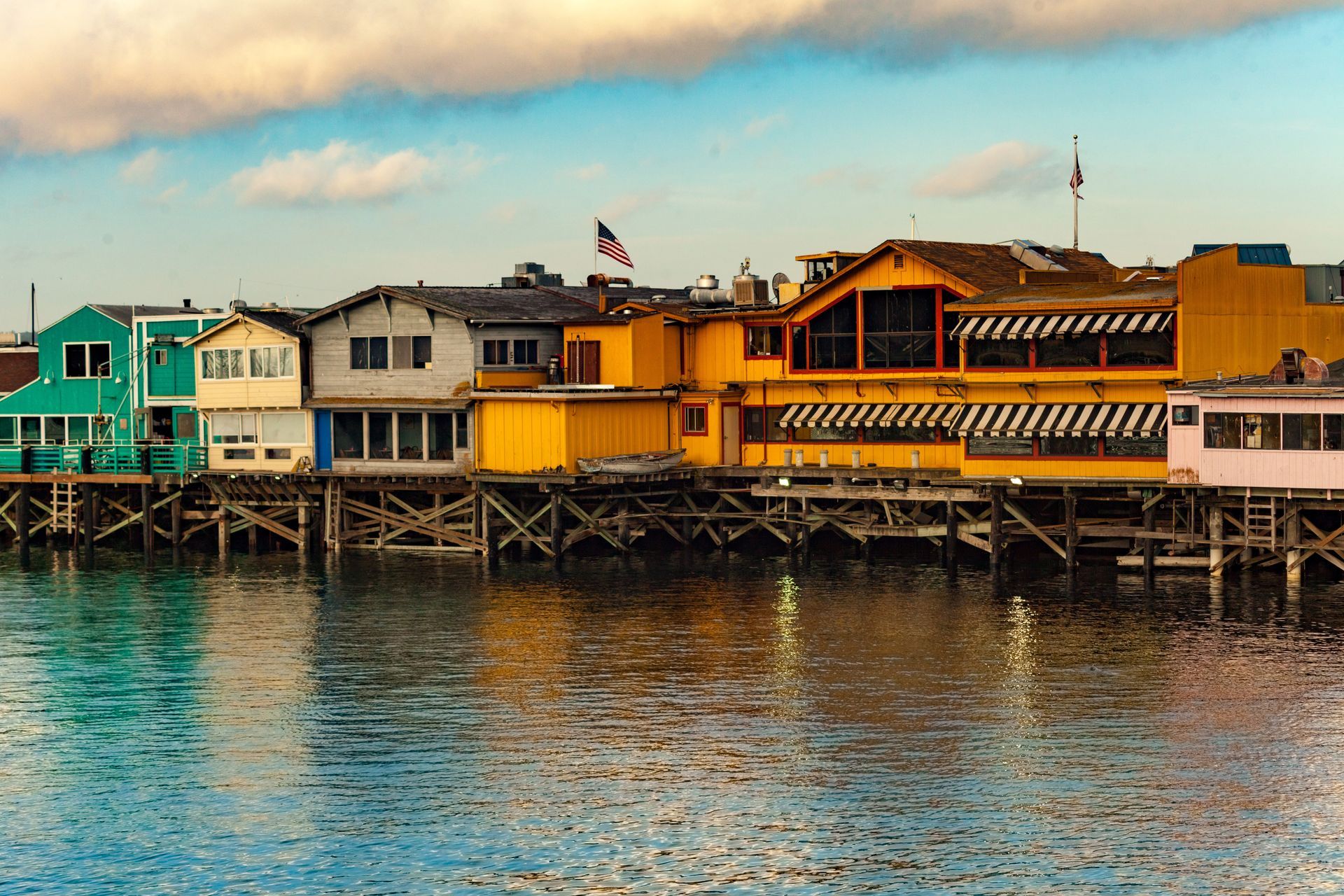 A line of colorful buildings on wooden stilts over calm water under a cloudy sky.