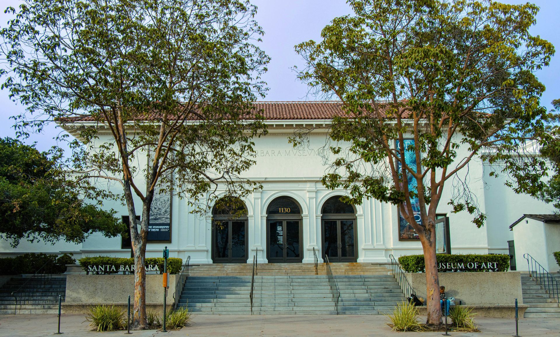 A white museum building with three arched doorways, tile roof, and two large trees in front under a clear sky.