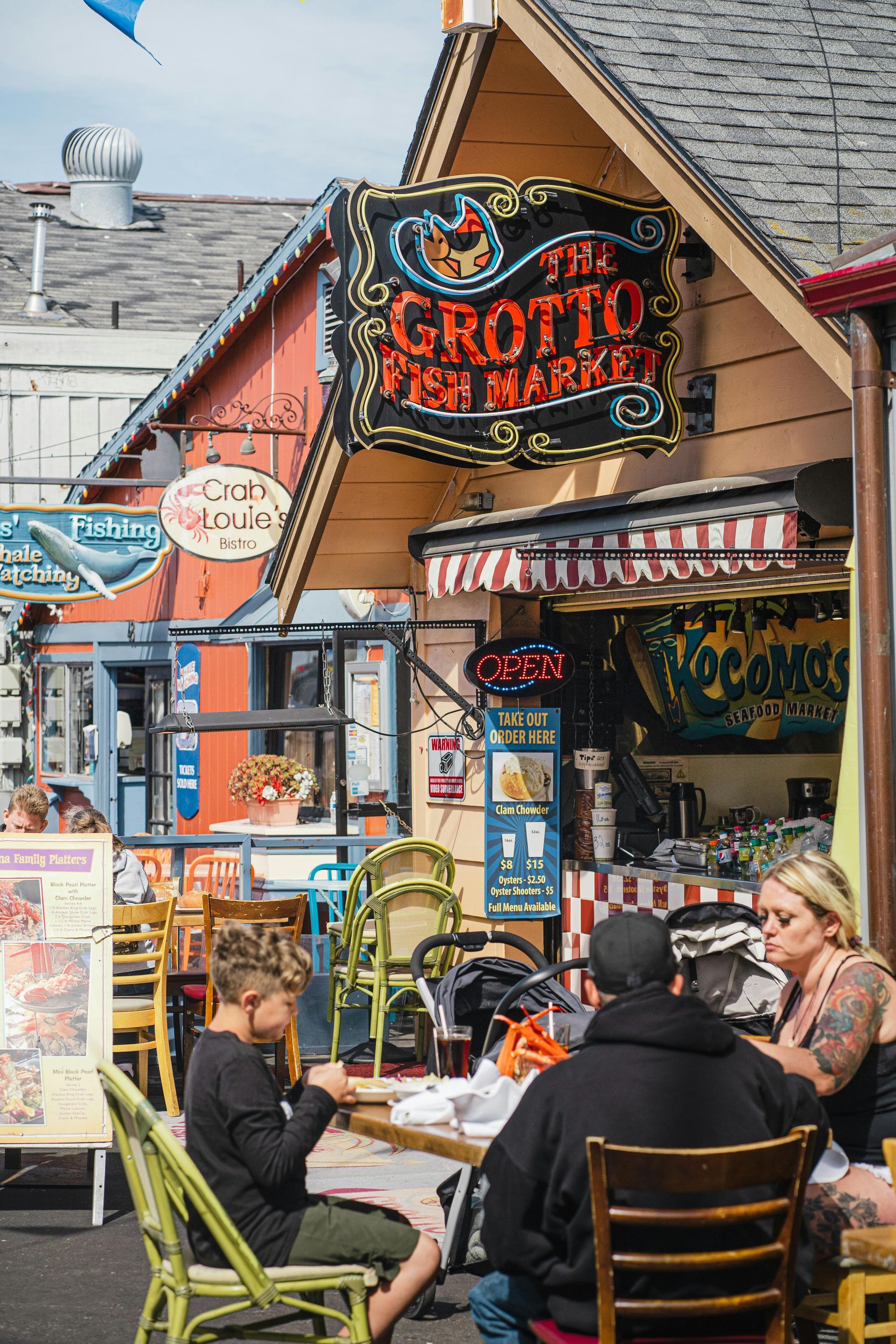 People dine at tables outside The Grotto Fish Market, a wooden-walled building with a black and red painted sign.