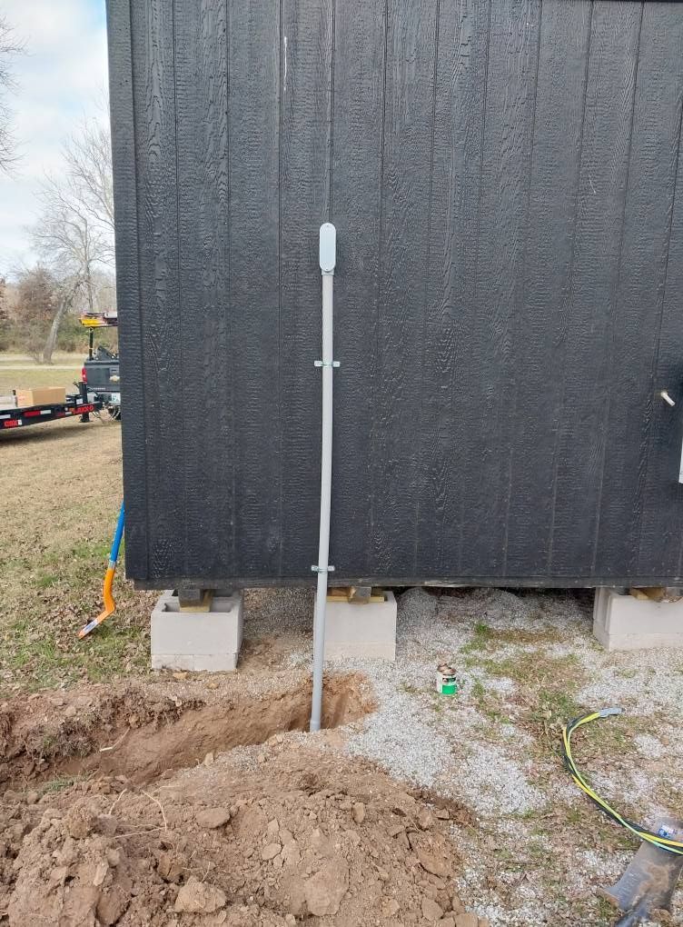 Grey conduit pipe installed beside a black building wall in a dirt trench near the foundation.