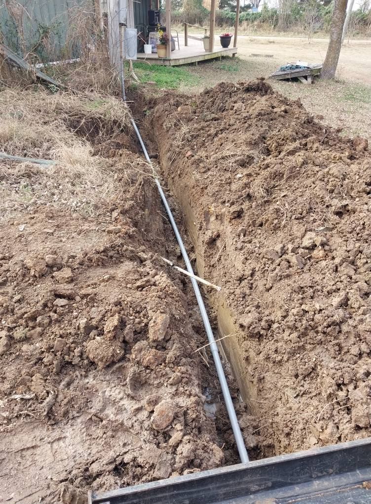 Trench with a black pipe laid in it beside a dirt yard and a small structure.