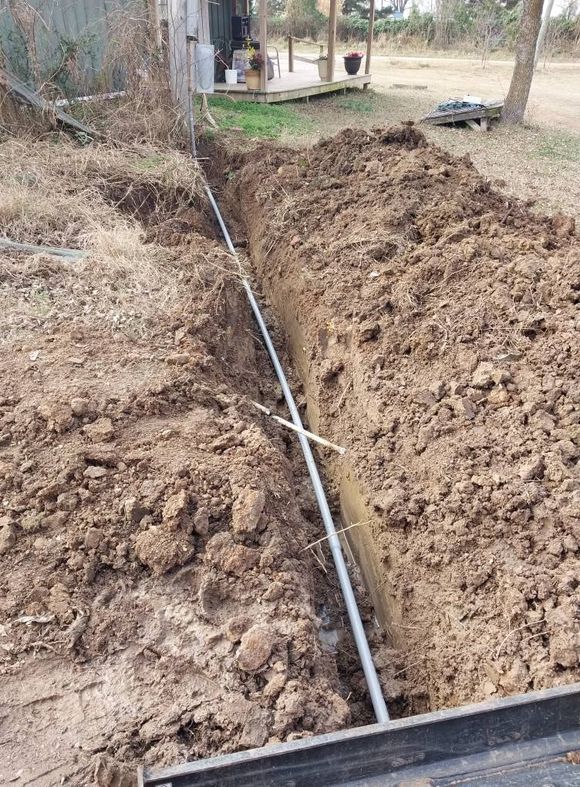 Trench with a black pipe laid along the bottom beside a rural building and dirt piles