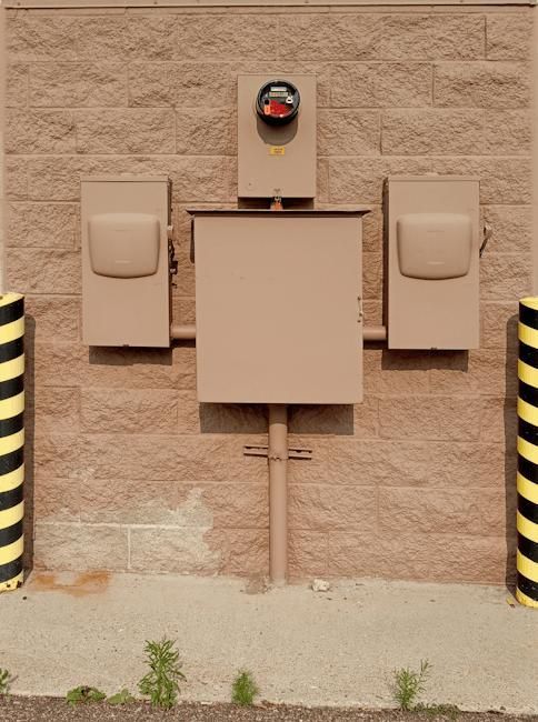 Wall-mounted utility box and electrical panels on a tan brick exterior wall, with yellow-black striped posts at sides