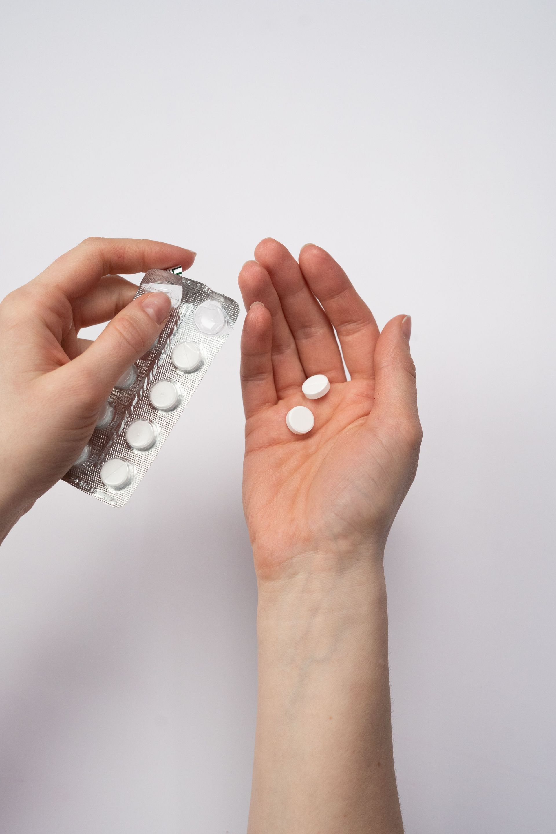 Hand holding two white tablets while another hand pours more from a blister pack on a white background