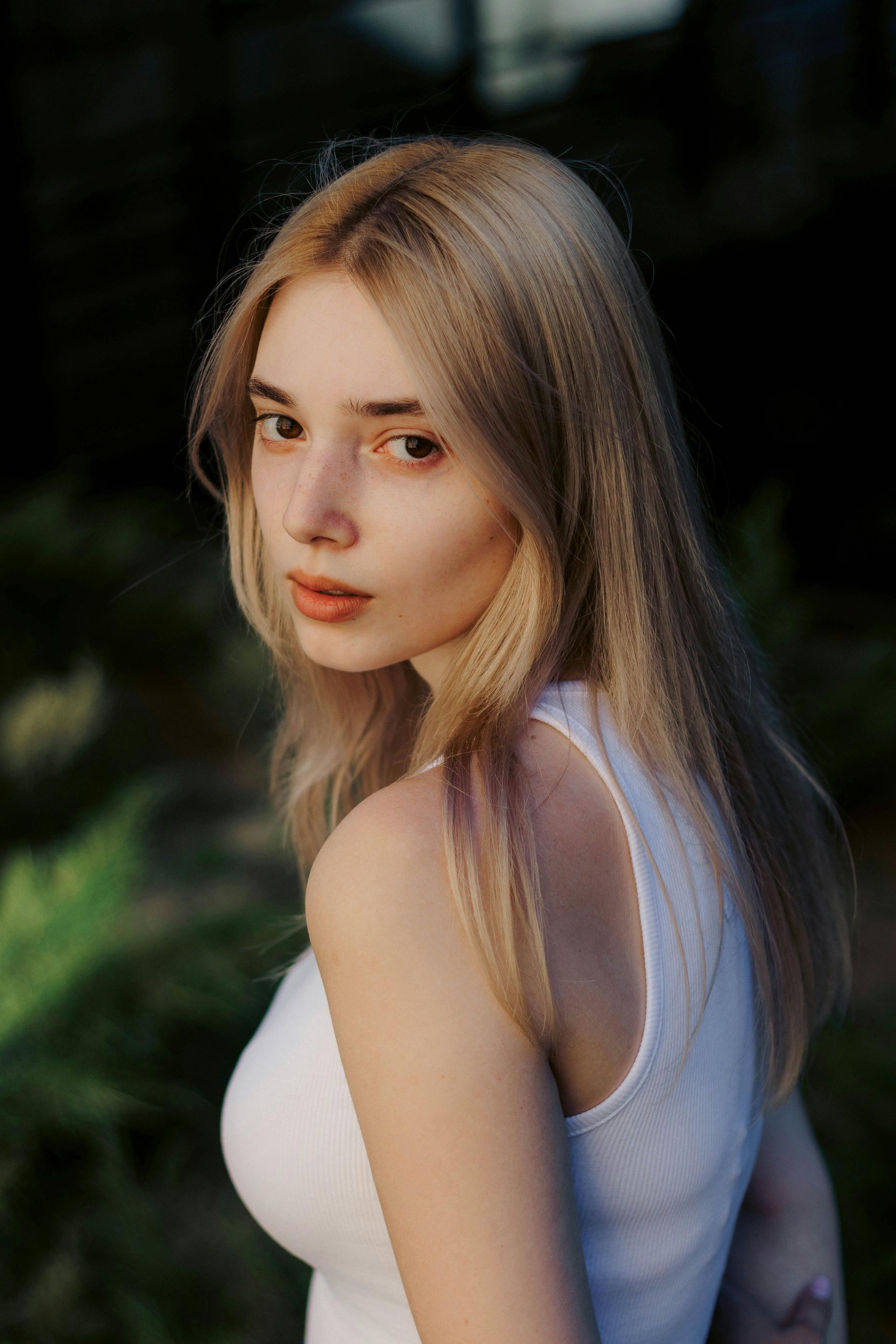 Vrouw met lang blond haar in een witte top, die over haar schouder kijkt tegen een donkere achtergrond.