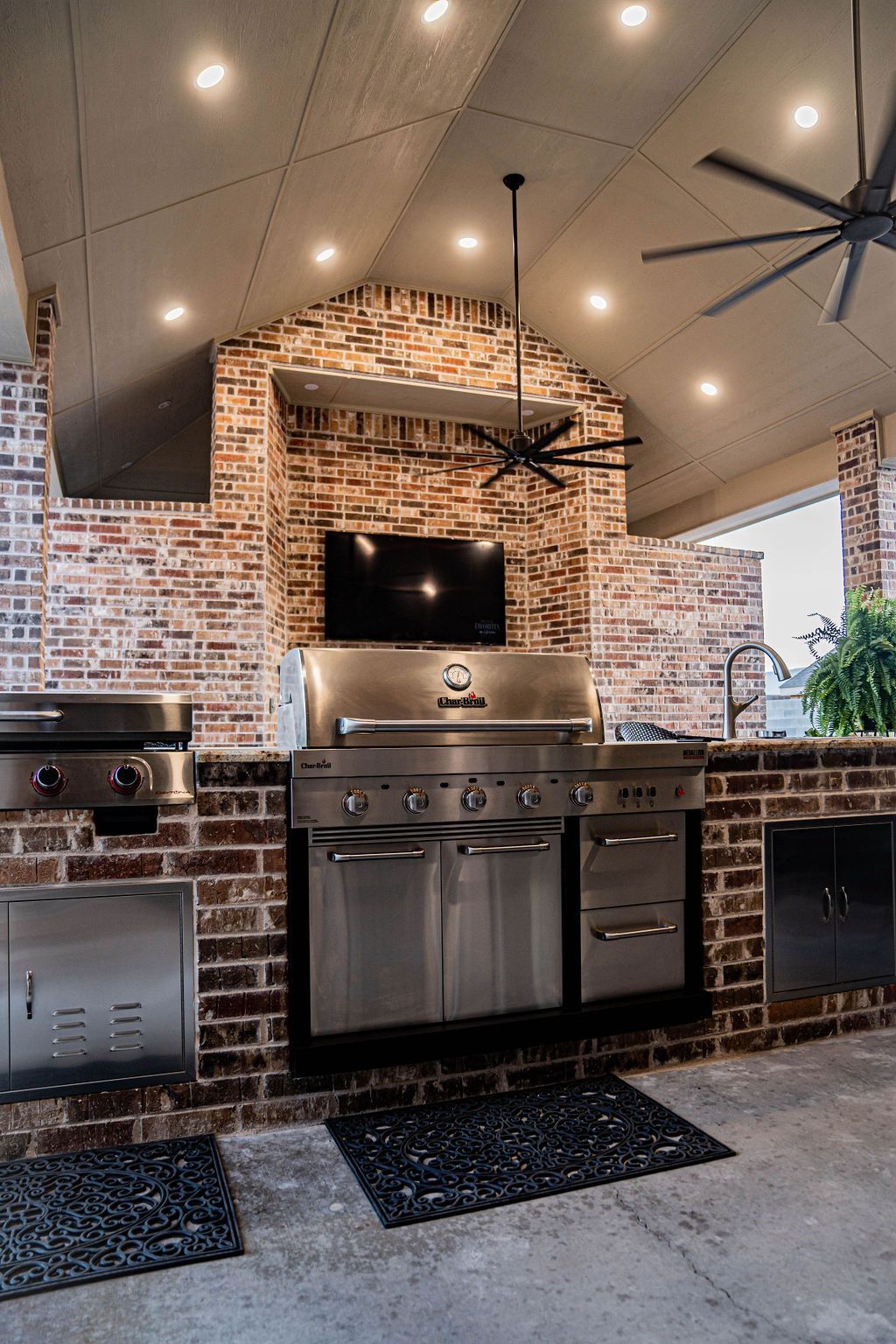 Outdoor kitchen with brick, stainless steel grill, TV, and ceiling fans.