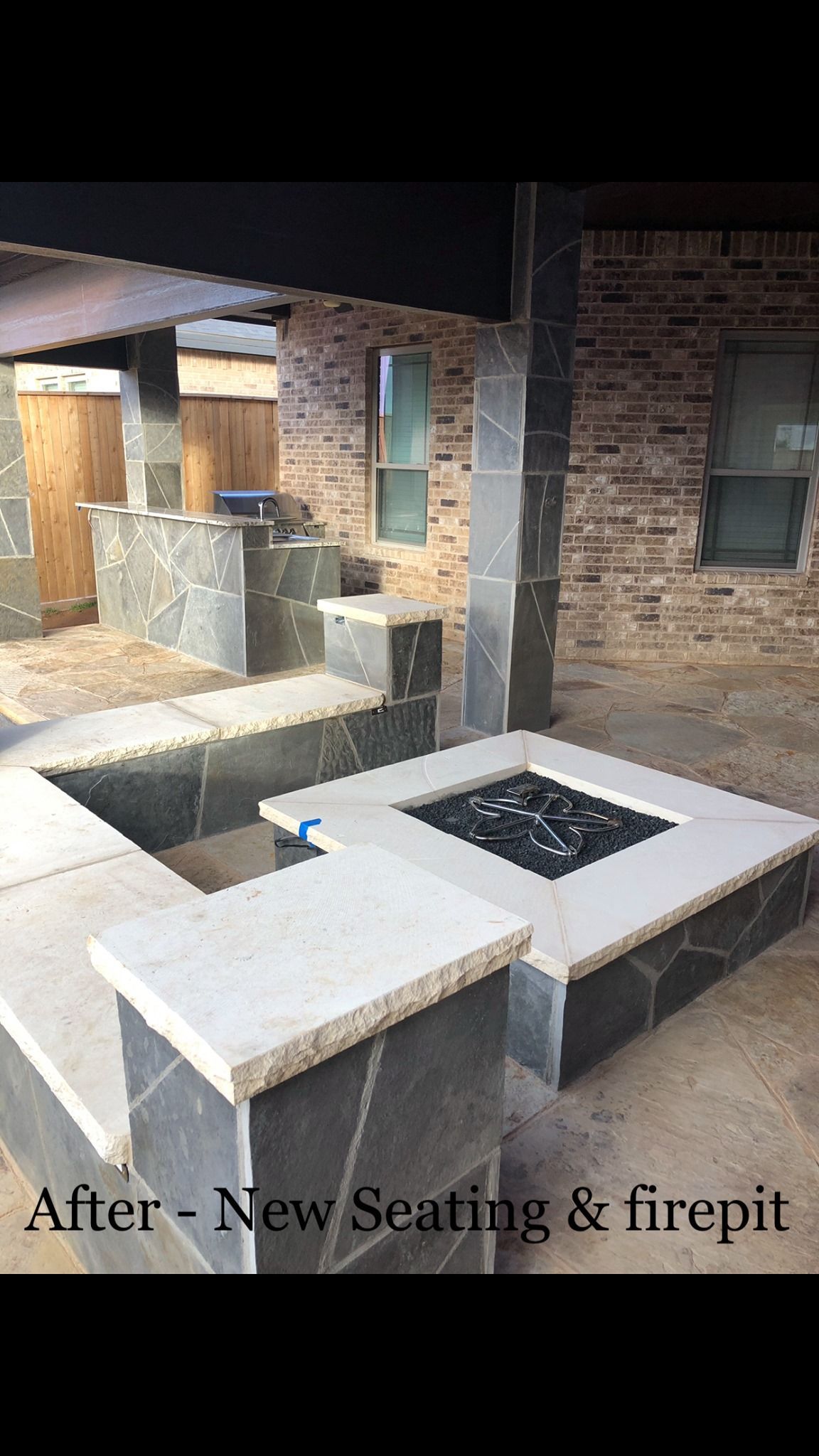 New patio with built-in seating and square fire pit; gray stone and black rocks. Brick wall in background.