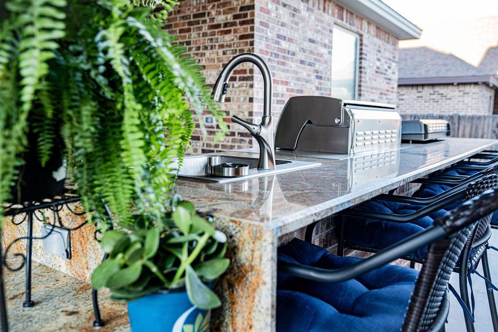 Outdoor kitchen with granite countertop, built-in grill, sink, and bar seating. Fern and potted plants.