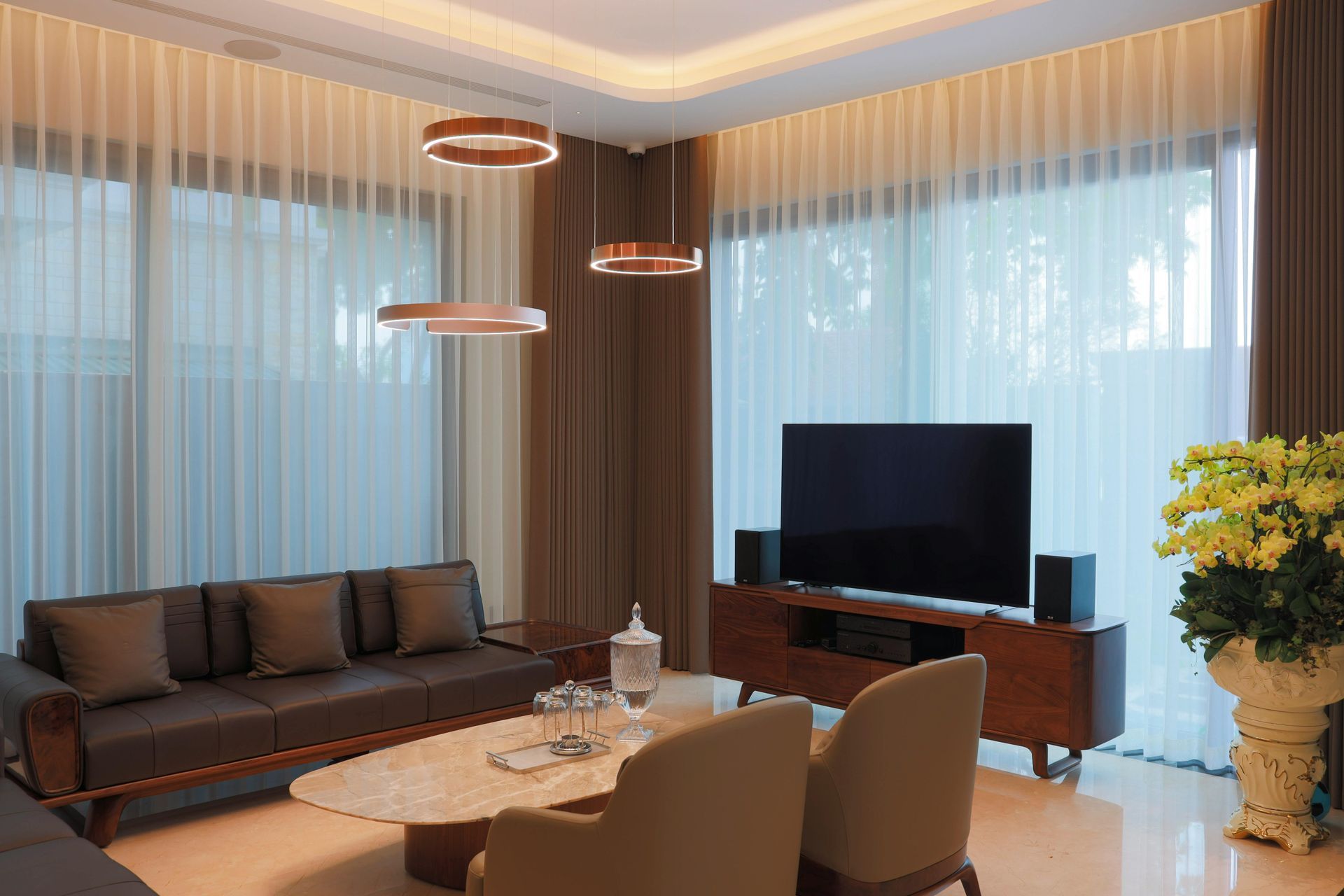 Modern living room with brown sofa, TV, and sheer curtains. Two hanging lights.