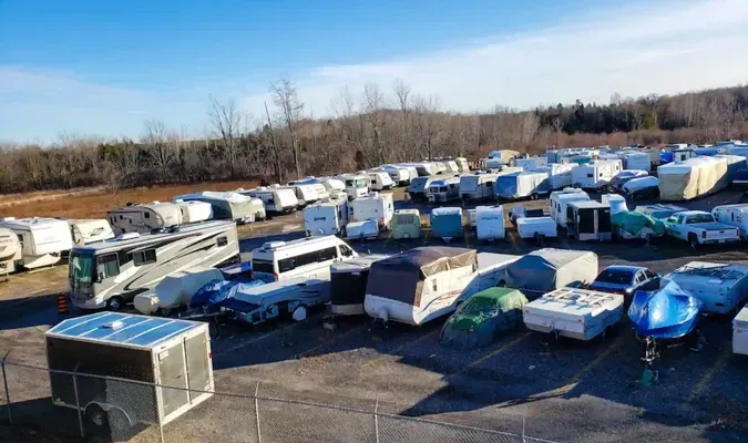 Rows of abandoned RVs and trailers in a snowy lot beside bare trees under a clear sky.