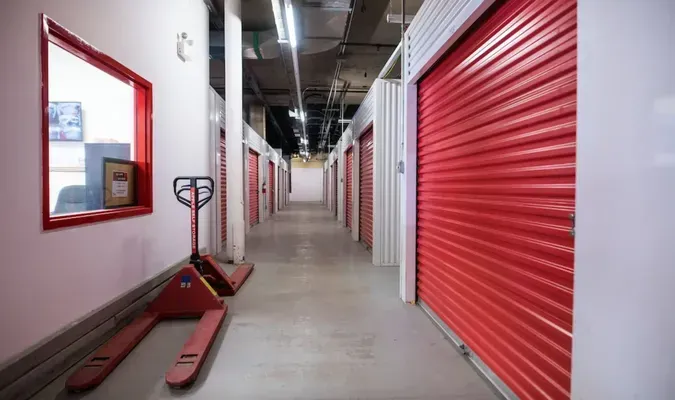 Storage facility hallway with red roll-up doors and a red pallet jack in the foreground