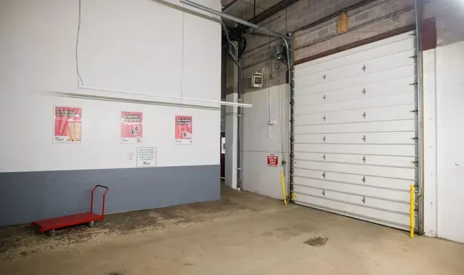 Empty warehouse corner with a large white roll-up door, concrete floor, and wall-mounted signs.