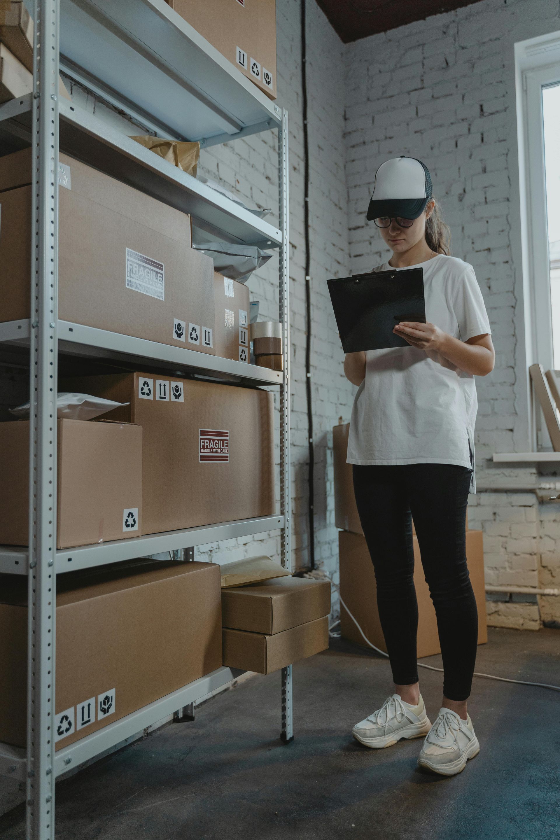 Person in white hard hat reading a clipboard beside warehouse shelves and stacked boxes