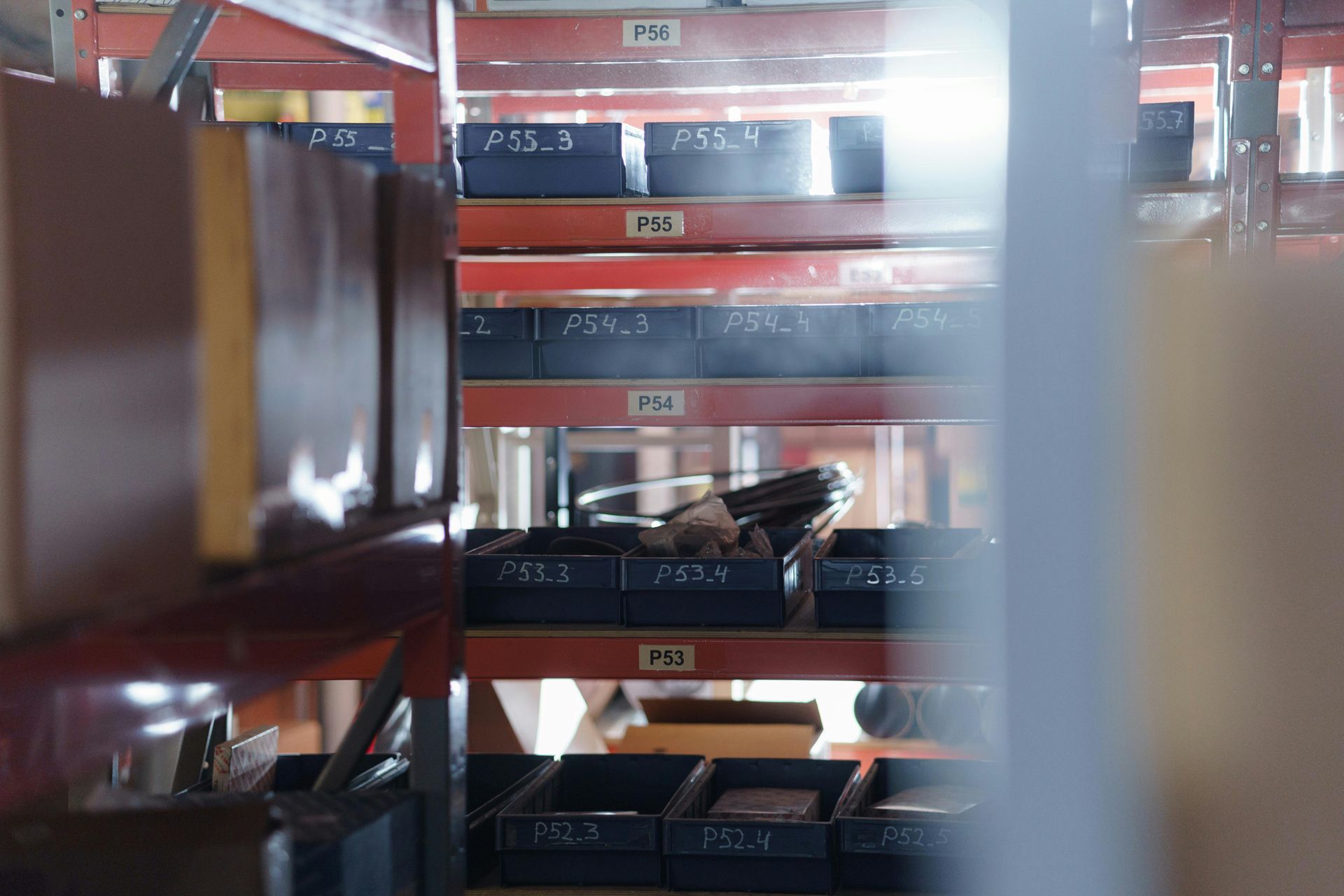 Red metal warehouse shelves with boxes and packages, seen through a narrow aisle.