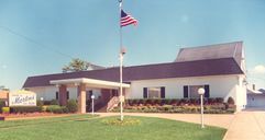 A wide, single-story building with a white exterior, dark roof, and covered entrance, with an American flag on a pole.
