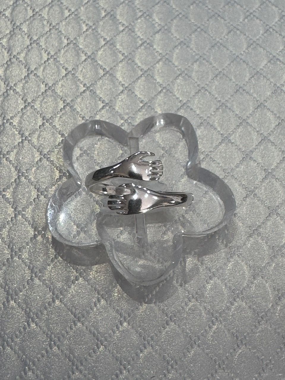 Anillo de plata sobre una exhibición transparente en forma de flor contra una superficie blanca texturizada.