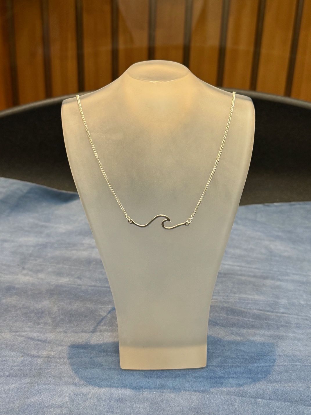 Collar colgante de ola de plata sobre busto de exhibición, sobre una superficie azul y un fondo de madera.