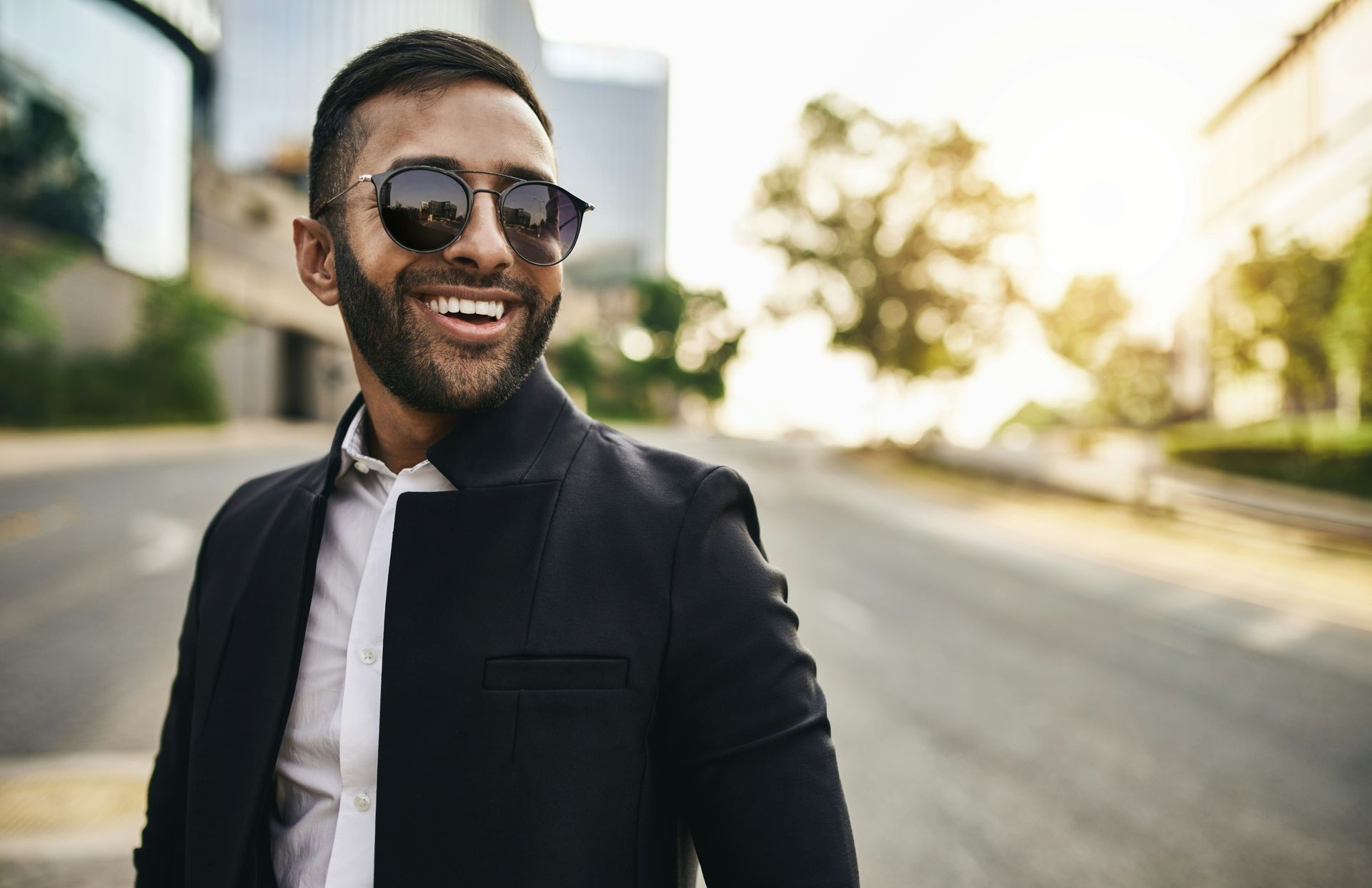 A man with a beard wearing sunglasses and a black jacket is smiling.