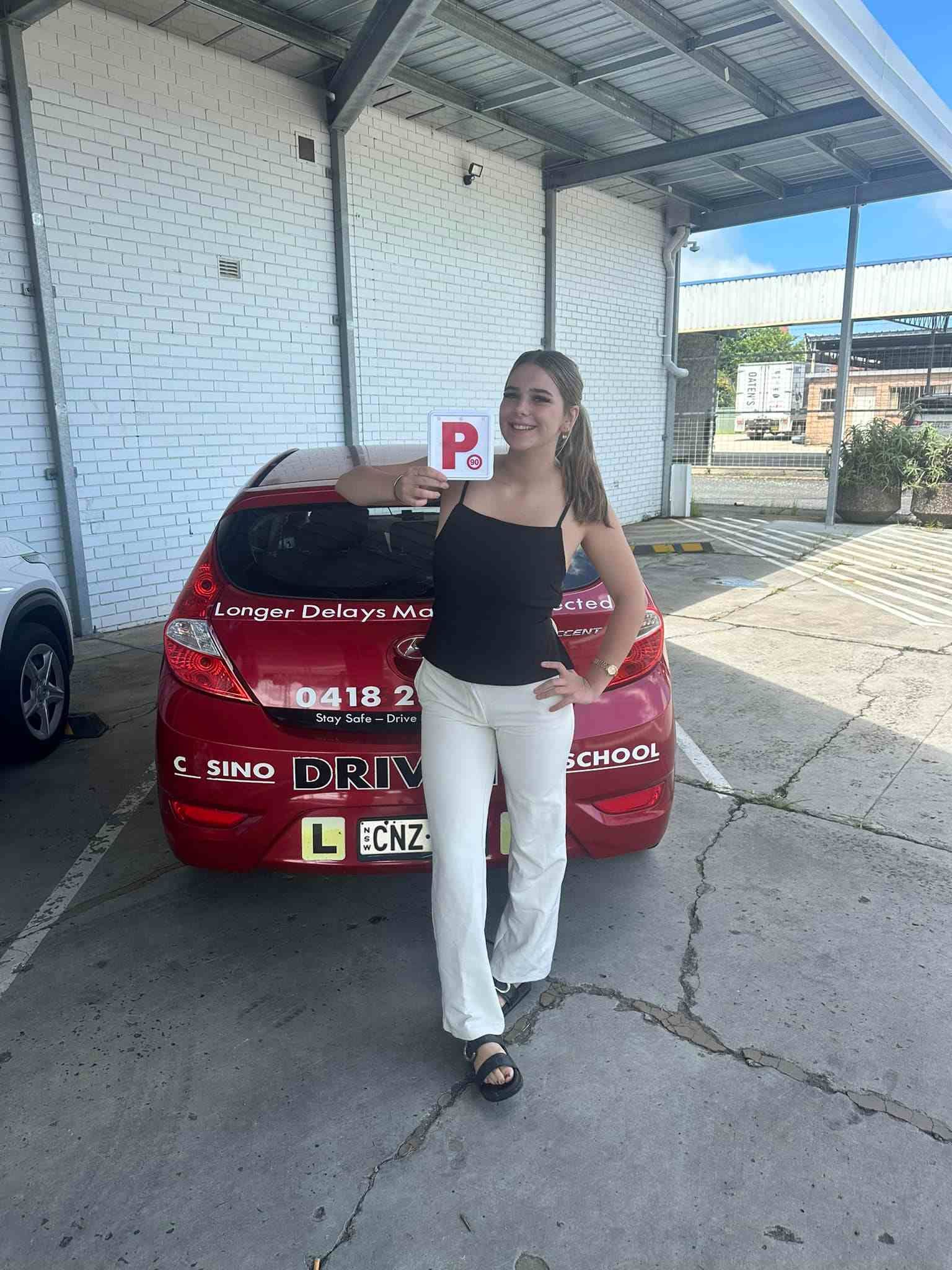 A Woman is Standing in Front of a Red Car Holding a R Sign — Casino Driving School In Ballina, NSW
