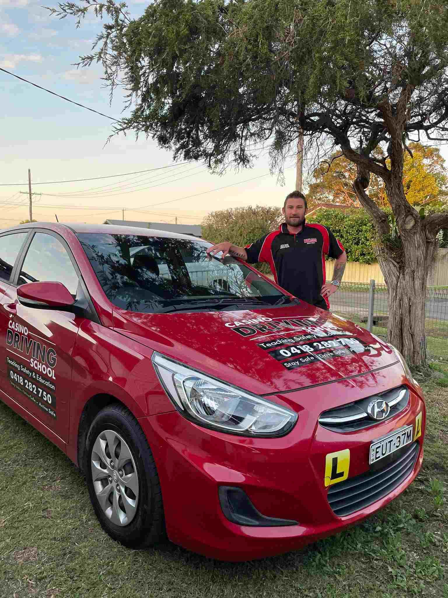 a Man is Standing Next to a Red Car in Front of a Tree — Casino Driving School In Casino, NSW