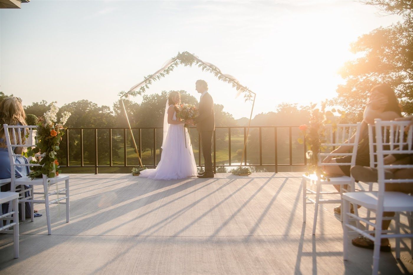 Bride and groom at outdoor wedding ceremony; gold sunlight, floral arch, white chairs, and guests.