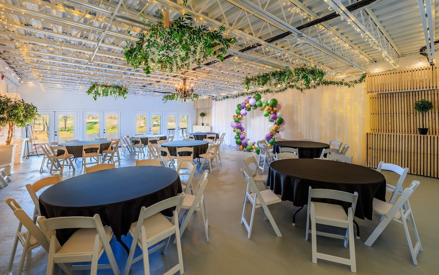 Event space with round tables, white chairs, and decorative greenery and balloons.