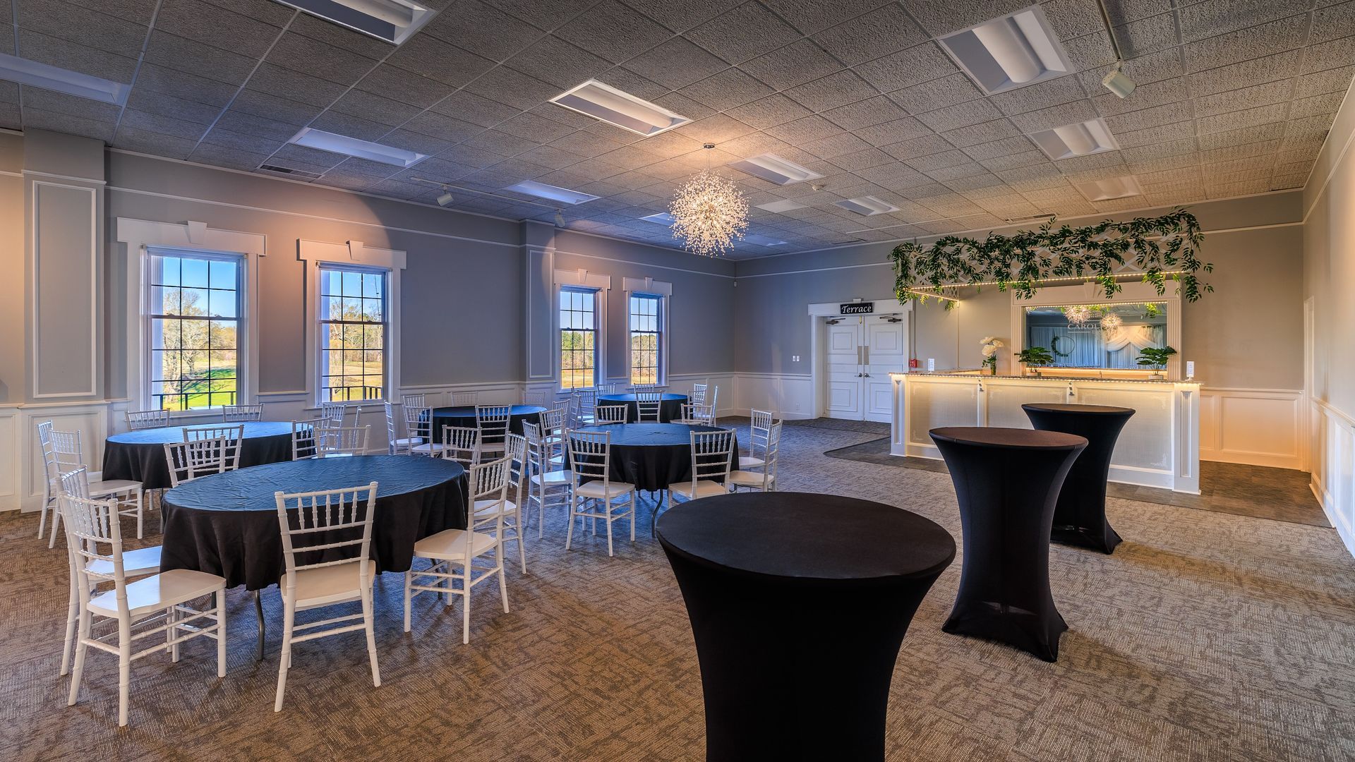 Event space with tables, chairs, bar area, and windows. Black and white color scheme.