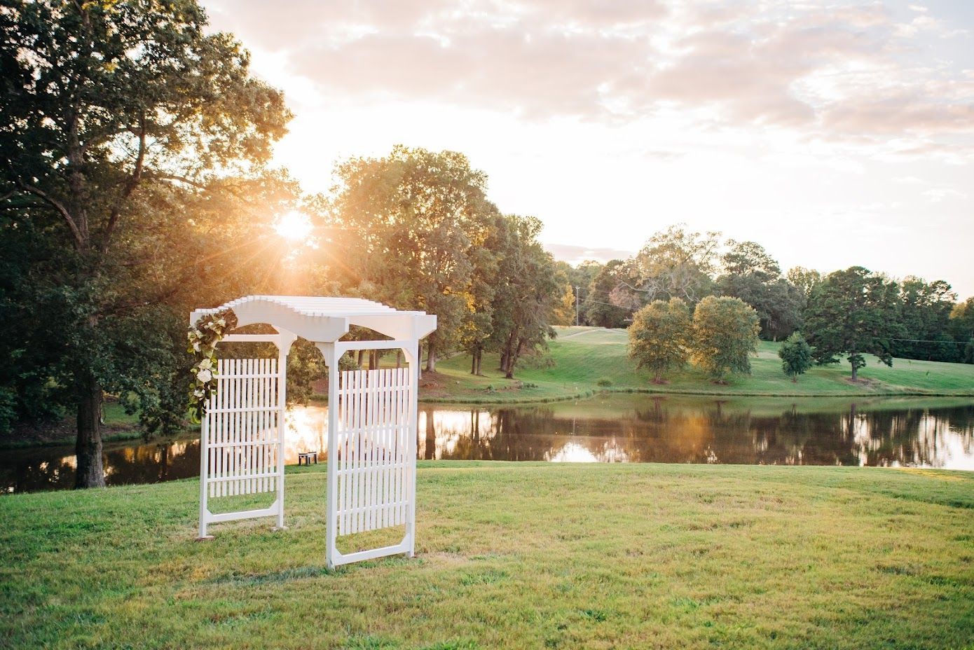 White arbor on a grassy lawn overlooking a lake at sunset.