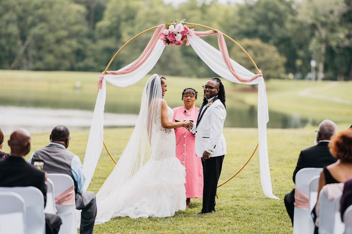 Wedding ceremony: two people under a floral arch exchange vows outdoors by a lake.