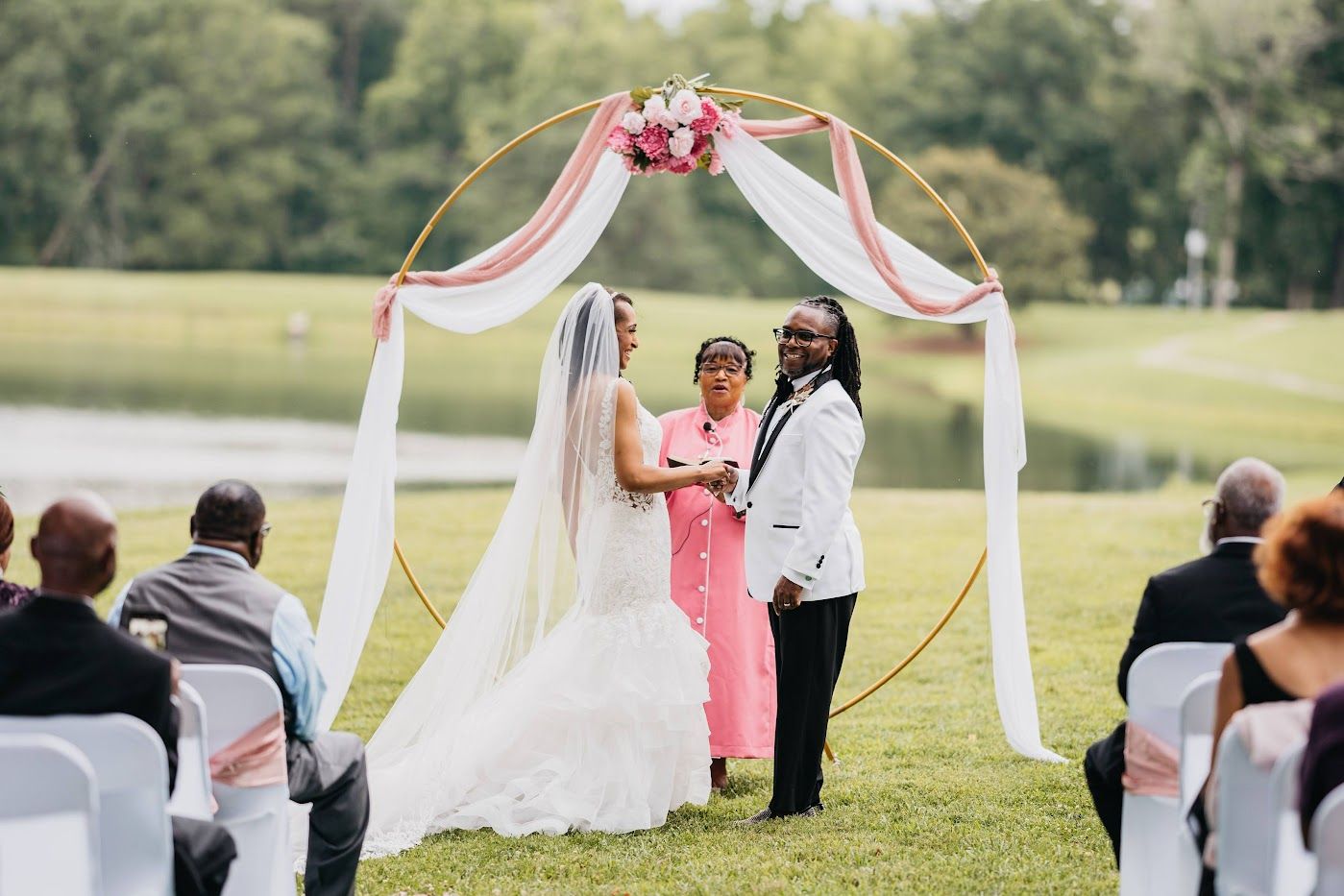 Wedding ceremony: two people under a floral arch exchange vows outdoors by a lake.