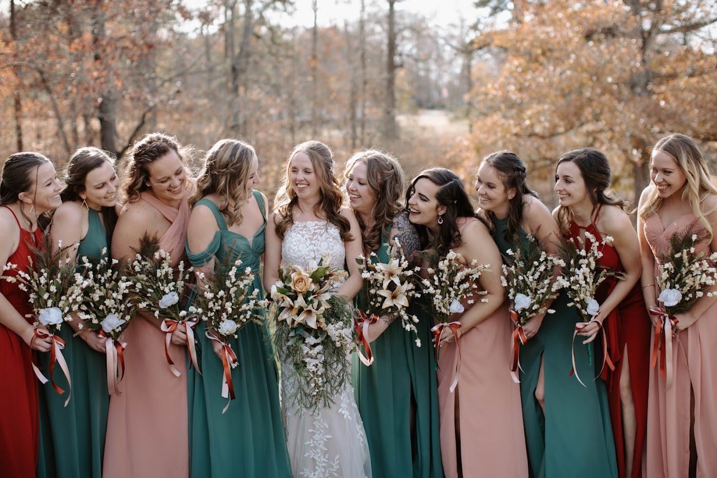 Bridesmaids in diverse dresses, and bride in white gown, smile and hold bouquets outdoors.