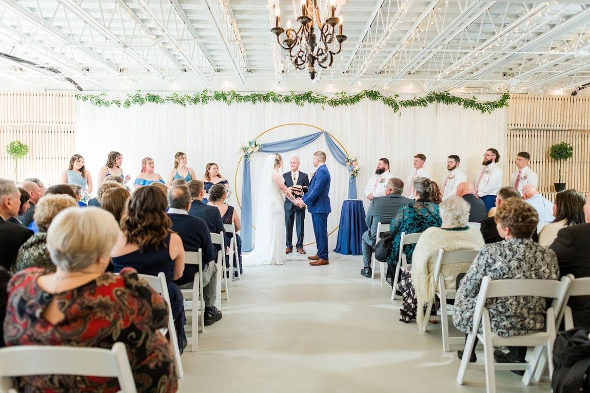Wedding ceremony indoors with bride, groom, wedding party, and guests seated in white chairs.