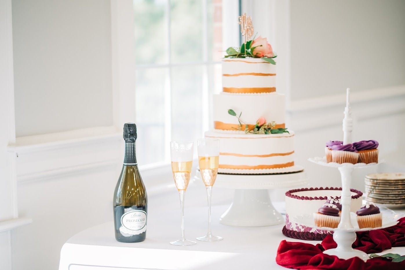 Wedding cake display with champagne bottle, glasses, cupcakes, and red cloth.