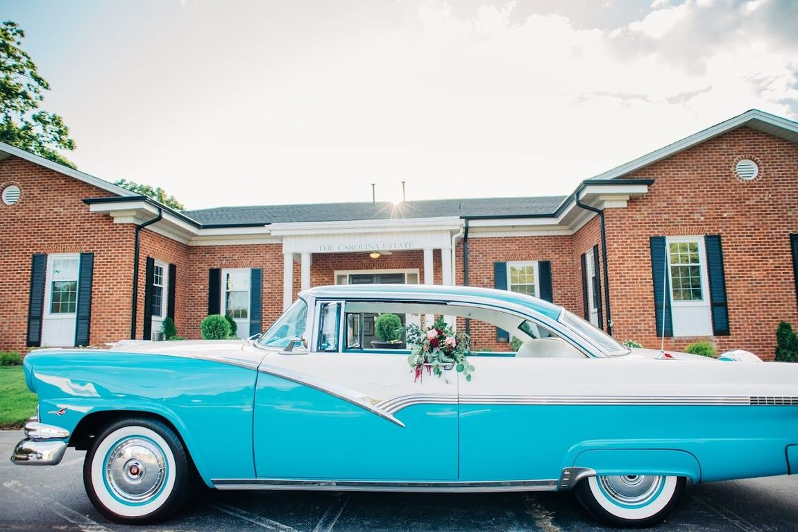 Turquoise and white vintage car parked in front of a brick building. Flowers on the car.