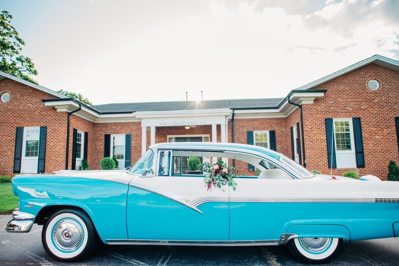 Turquoise and white vintage car parked in front of a brick building. Flowers on the car.