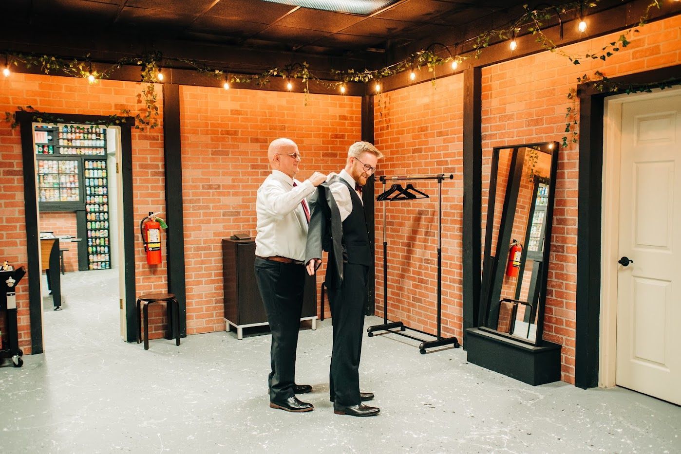 Man being helped with his suit by another man in a room with brick walls, a mirror, and hanging lights.
