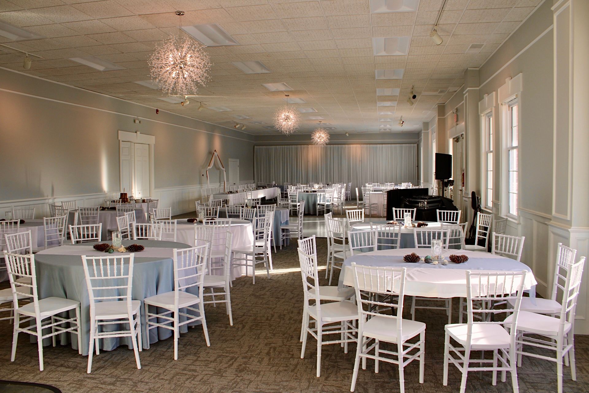 Interior of a banquet hall with round tables set for a formal event. White chairs, light blue tablecloths, and chandeliers.
