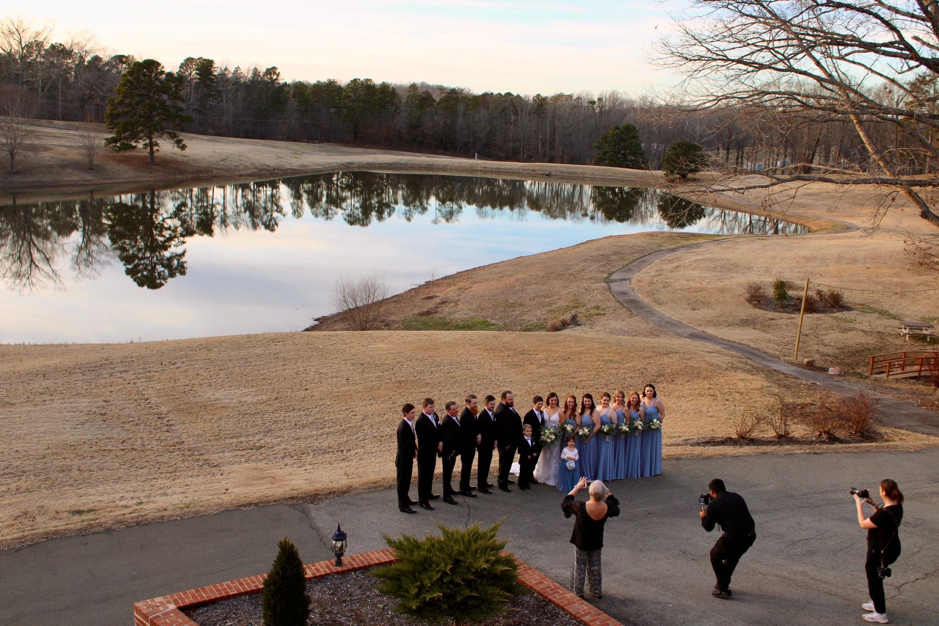 Wedding party poses by a lake; bridesmaids in blue, groomsmen in black. Photographers take photos.