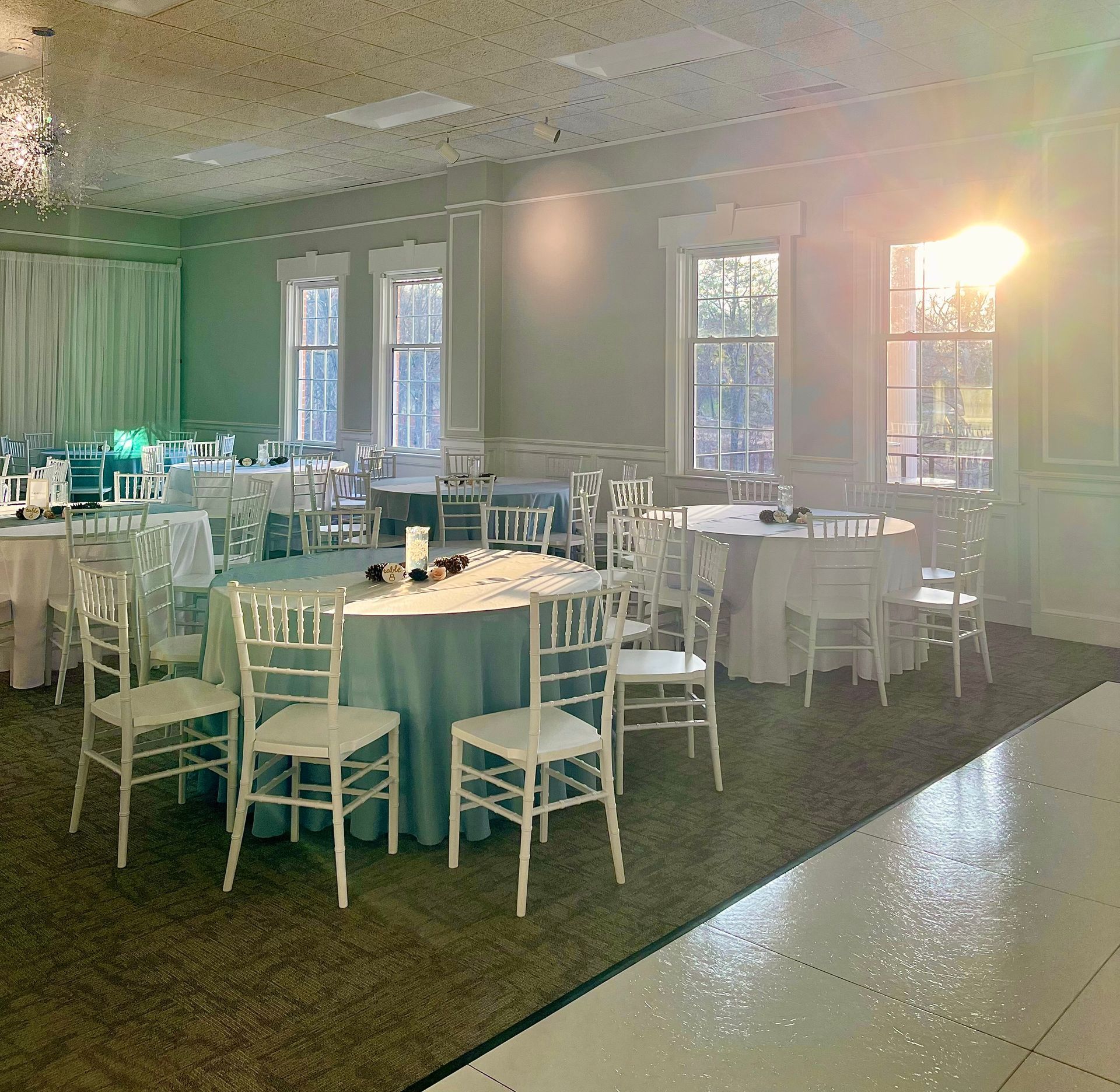 Reception hall with round tables set for a party; white chairs, blue tablecloths. Windows with sunlight.