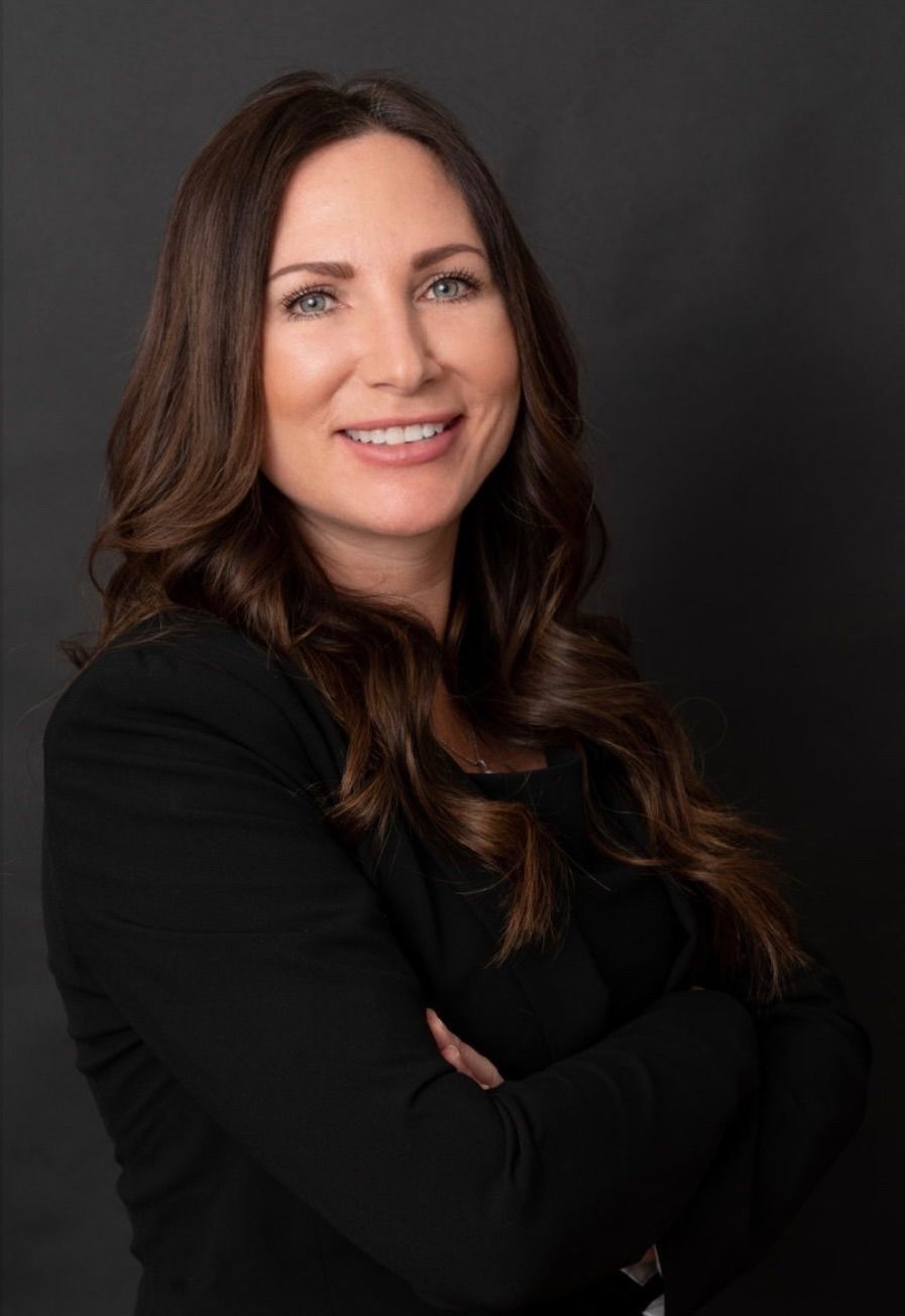 Maureen Roberts headshot. A woman smiling, with dark brown curly hair, wearing a black blazer and has her arms crossed.