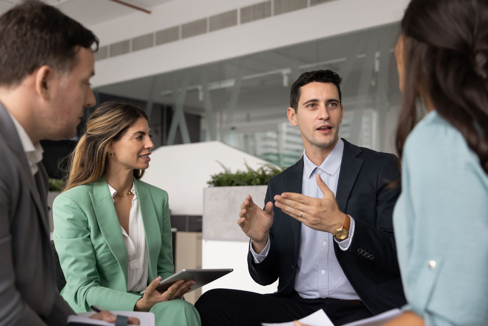 Colleagues in a meeting room discussing ideas, one man gesturing while others listen with a tablet