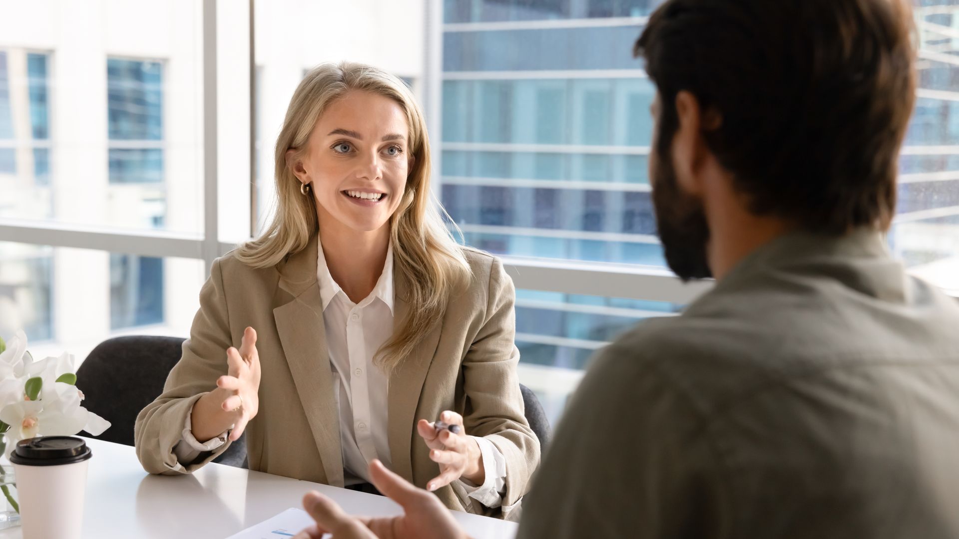 Two people in a bright office meeting, one woman speaking and gesturing across a table.