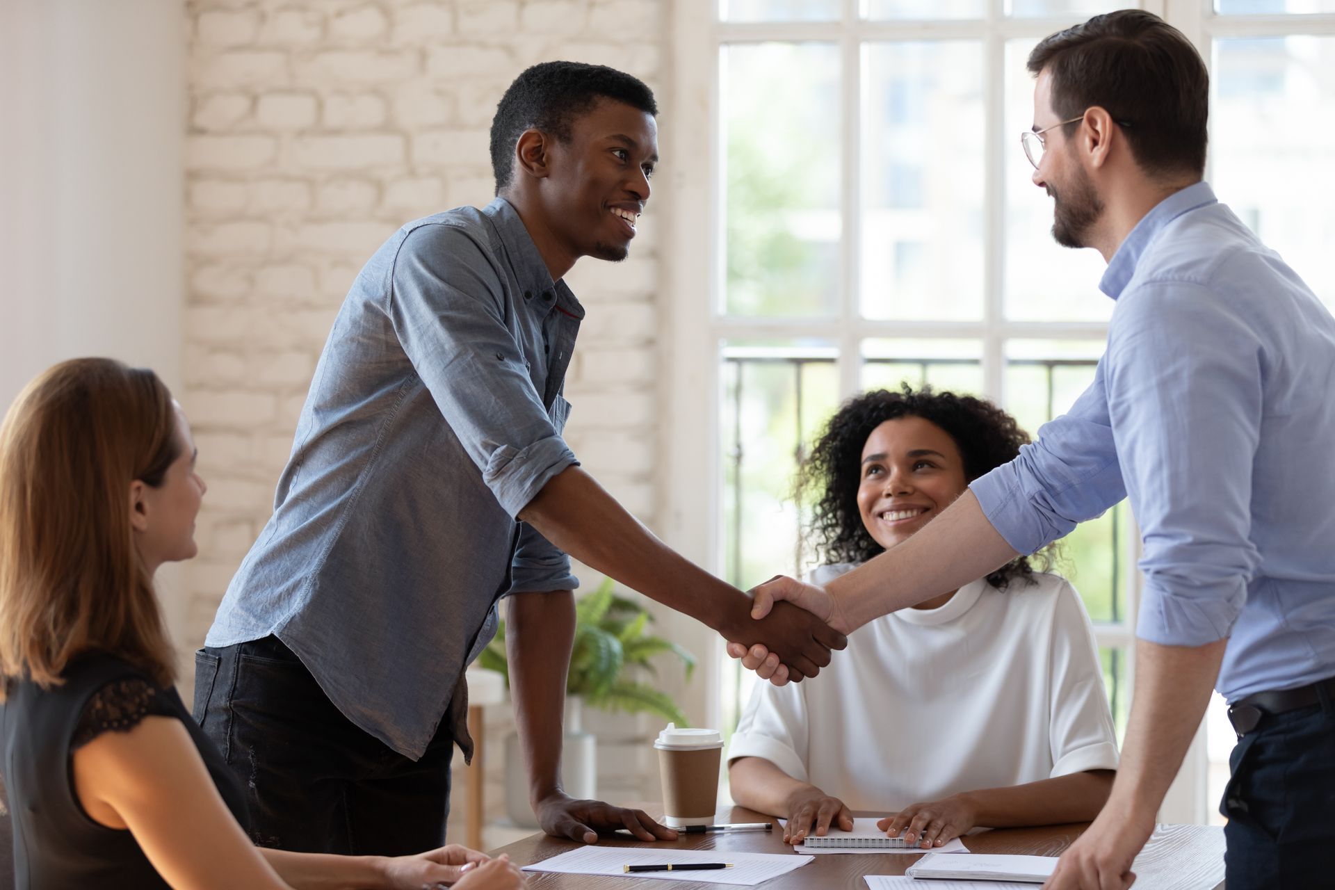 Two coworkers shake hands across a meeting table in a bright office conference room.