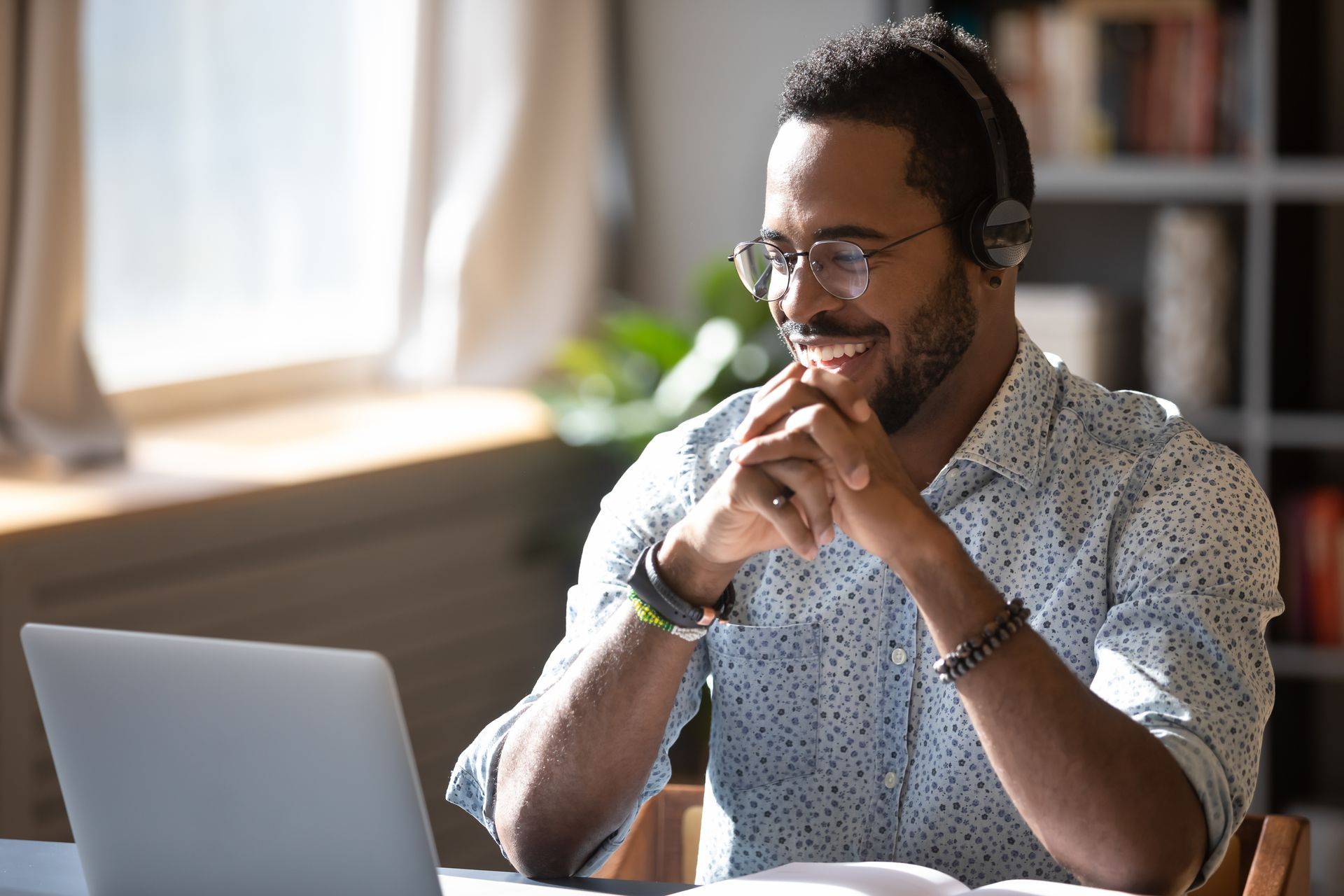 Smiling person with glasses working on a laptop at a table in a sunlit room