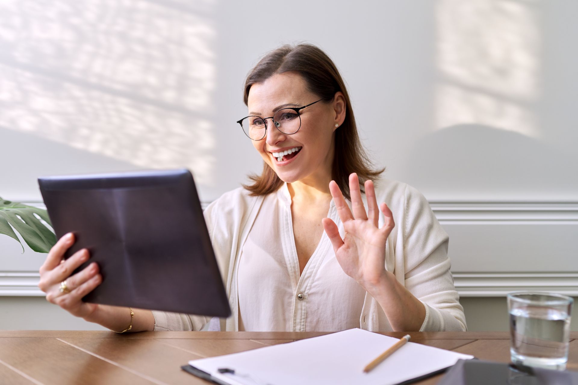 Woman on a video call, smiling and waving at a tablet in a bright office with a notebook and glass of water.